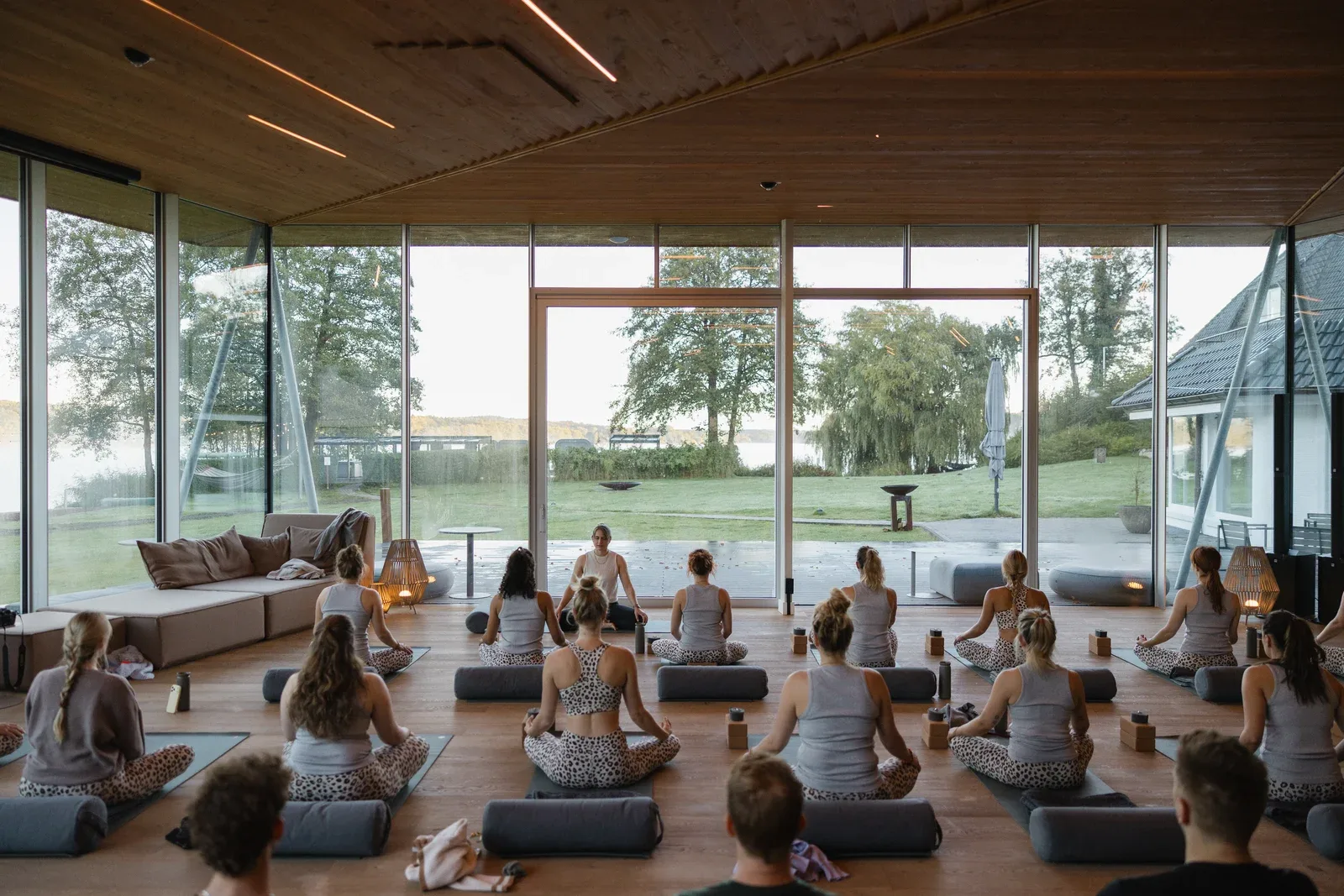 Yogagruppe auf Matten im Raum mit Glasfront, Blick über die Seewiese auf den Bistensee am Morgen
