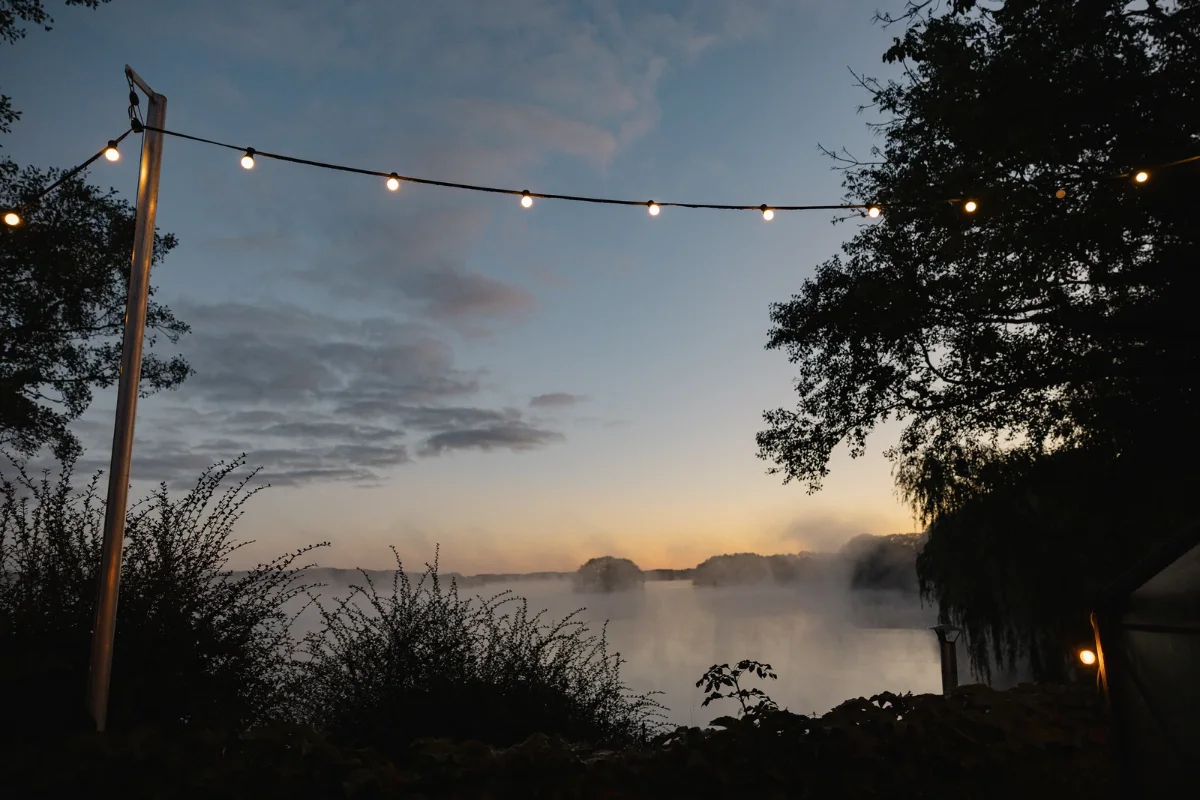 Morgennebel über dem Bistensee, davor eine Lichterkette in der Dämmerung