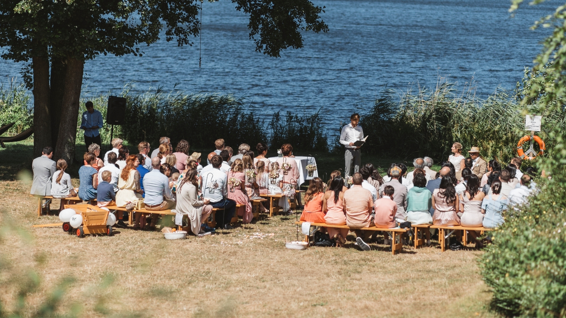 Freie Trauung mit vielen Gästen auf Holzbänken am Ufer eines Sees. - Seehotel Töpferhaus in Schleswig-Holstein