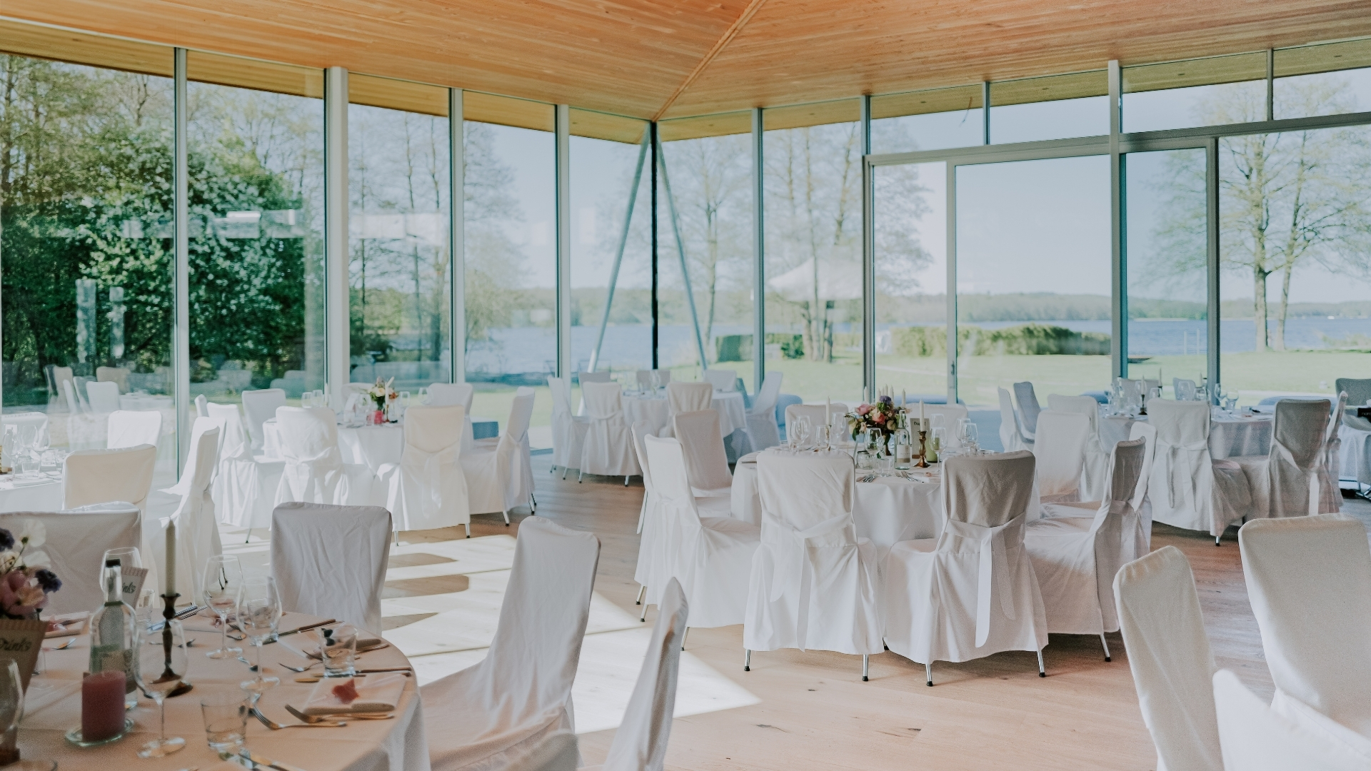 Festlich gedeckte Tische mit weißen Hussen in einem lichtdurchfluteten, modernen Raum mit Panoramablick auf Natur und See - Seehotel Töpferhaus in Schleswig-Holstein