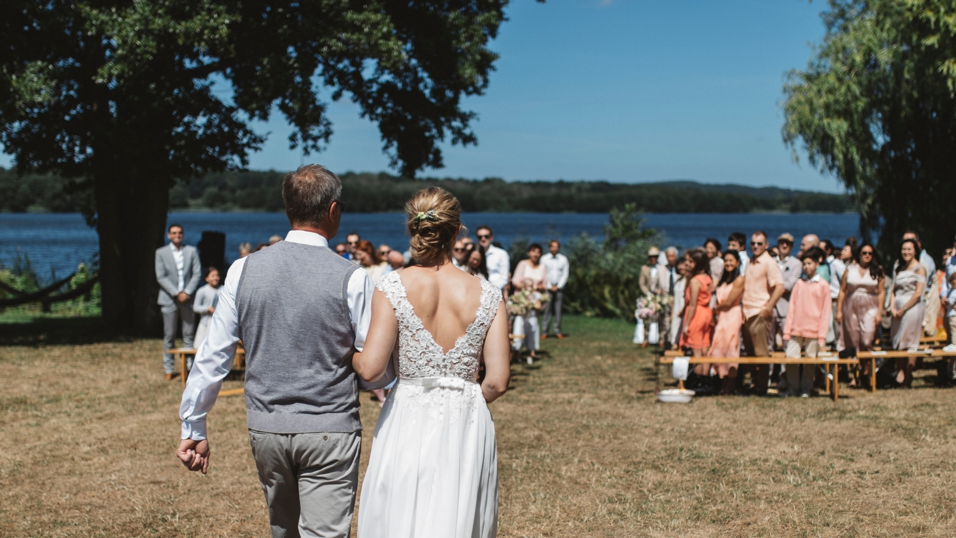 Braut und ihr Vater gehen bei sonnigem Wetter auf einer Wiese am See Richtung Hochzeitsgesellschaft. - Seehotel Töpferhaus in Schleswig-Holstein